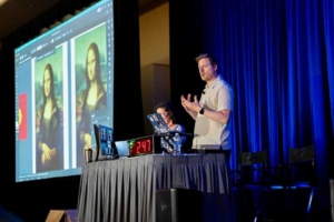 Presenter on a stage gesturing toward a large screen displaying a photo-editing app with two Mona Lisa images, two laptops on a table, and a blue curtain backdrop.