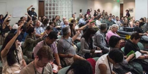 Large audience in a conference room, many people seated and raising hands as if asking a question or voting.