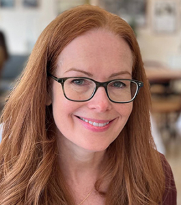 Smiling woman with long red hair and rectangular glasses in a bright indoor setting, facing the camera.