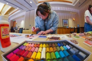 Person working on a craft project at a table with colorful pastels and supplies in a workshop setting, foreground shows a tray of bright colors.