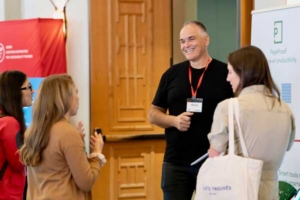 Smiling man with a lanyard talks with three attendees at a conference booth by a red banner and display stands.