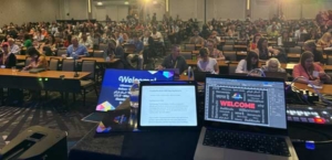 Audience seated in a large conference hall watching a stage with two laptops displaying a welcome screen.