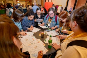 A diverse group of people gathered around a round table at a conference, examining maps and papers and taking notes together.
